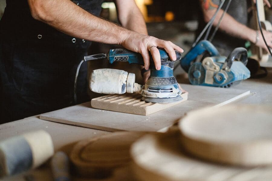 A carpenter using a random orbital sander to polish a wooden board at a workbench